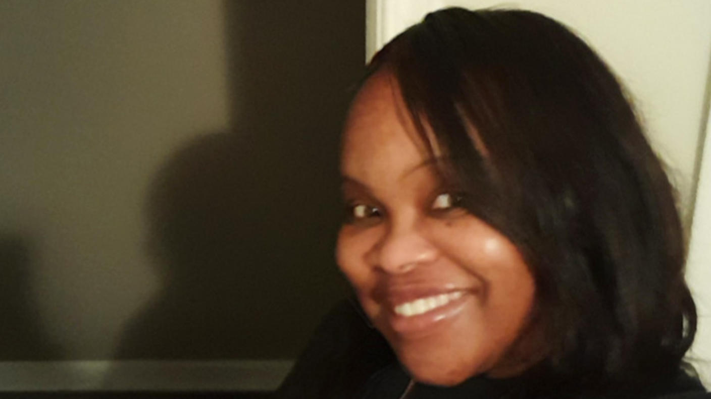 A black woman wearing a black t-shirt and chain necklace, taking a selfie in her home.