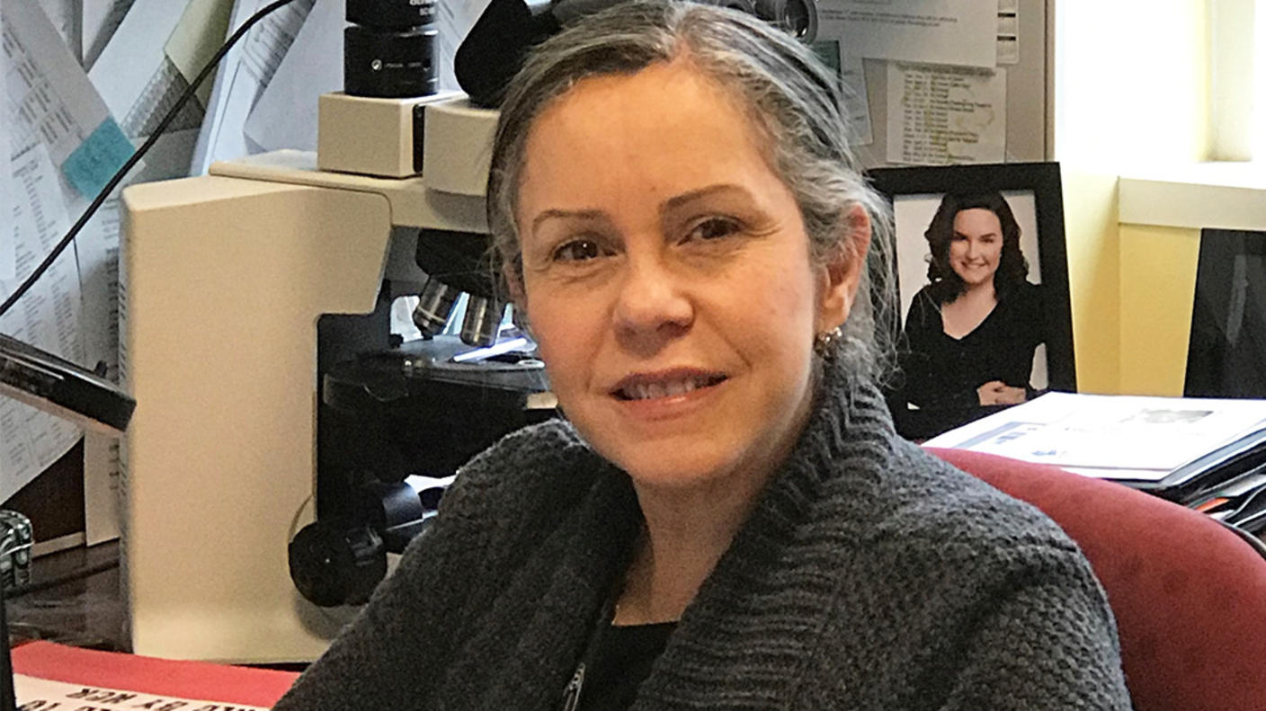 Puerto Rican woman (Nilsa Ramirez) sits at a desk with a microscope, framed photo, and papers tacked to a bulletin board behind her.