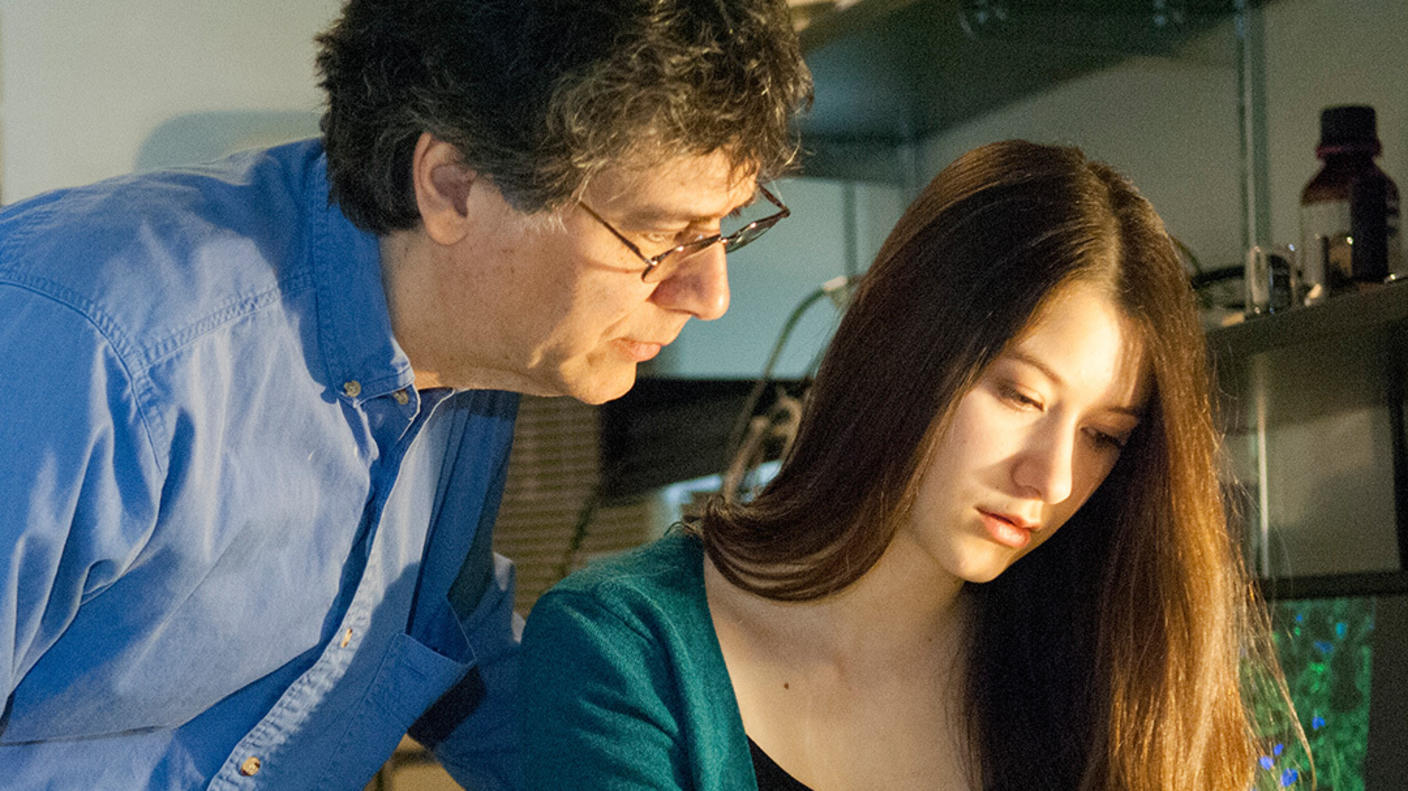 A young woman sits looking and pointing a pen at a paper on a desk in a lab office. Her father leans over her shoulder looking at the same paper. 