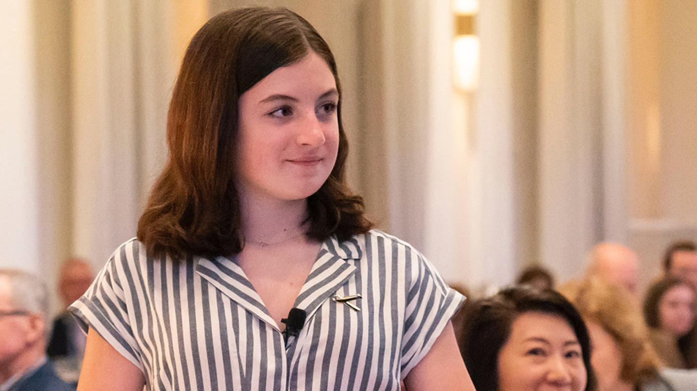 A young woman (Grace) with shoulder-length brown hair is standing and smiling in a room filled with seated people. She is wearing a striped dress with a tie at the waist and has a microphone clipped to her collar. In the background, attendees are seated at tables, engaging in conversation and looking towards the person standing.