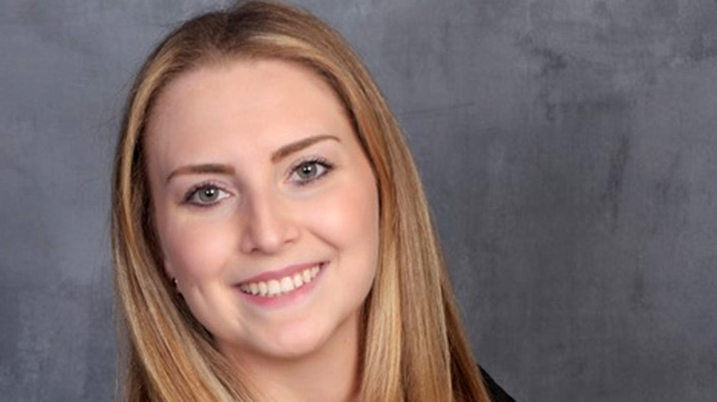 Woman with light skin and long, straight blonde hair wearing a pale pink blouse under a dark jacket stands smiling at the camera.