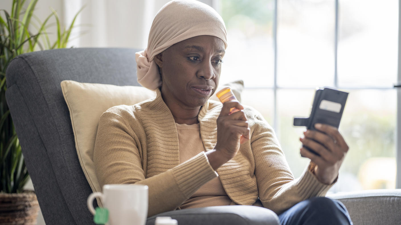 Woman wearing a head scarf holding a bottle of medication in one hand and looking at her phone in the other hand.