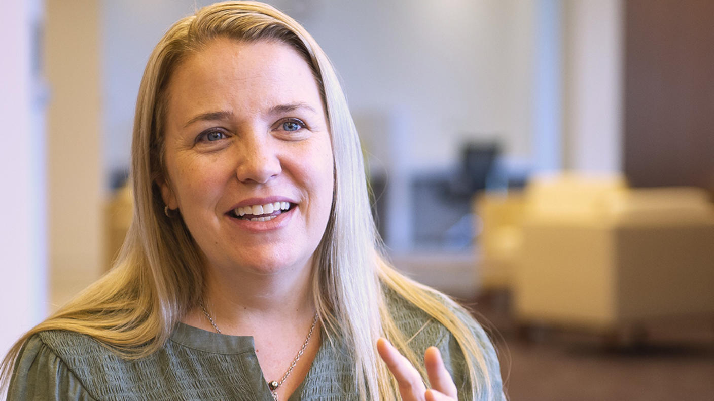 A woman (Dr. Emily Tonorezos) with long blond hair wears a muted green button-up blouse and sits in a chair, smiling and talking with her hands.