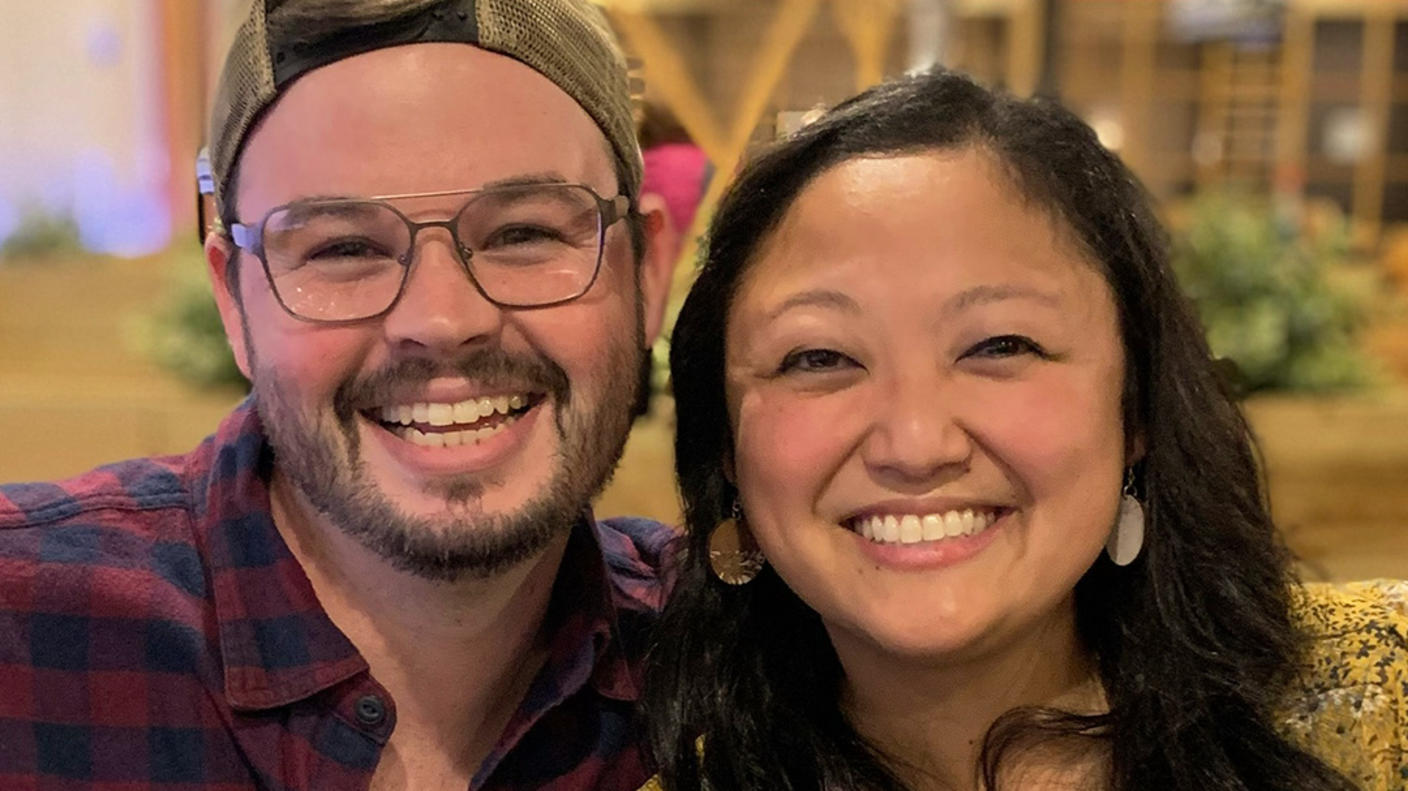 Woman (Sarah) with long black hair, earrings, and peach-colored blouse with blue and white flowers sits next to her husband. Both smile at the camera.