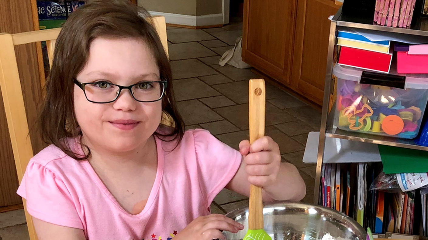 A girl, Abby, wearing glasses and a pink T-shirt, stirs ingredients in a metal bowl with a rubber spatula and smiles at the camera.