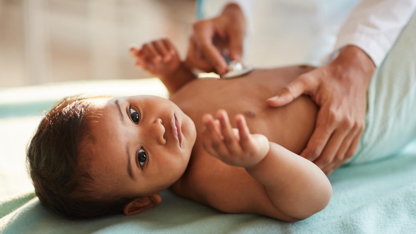 Photo of an infant lying on an exam table with an adult holding a stethoscope to the baby's chest.
