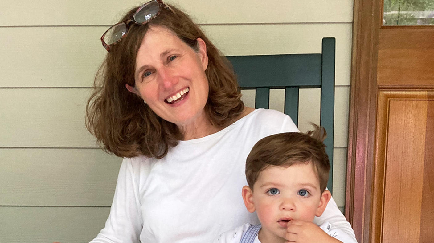 A smiling woman, Dr. Corinne Linardic, with red hair wearing a white shirt and a long, tan skirt sits in a chair with her arm around a child.