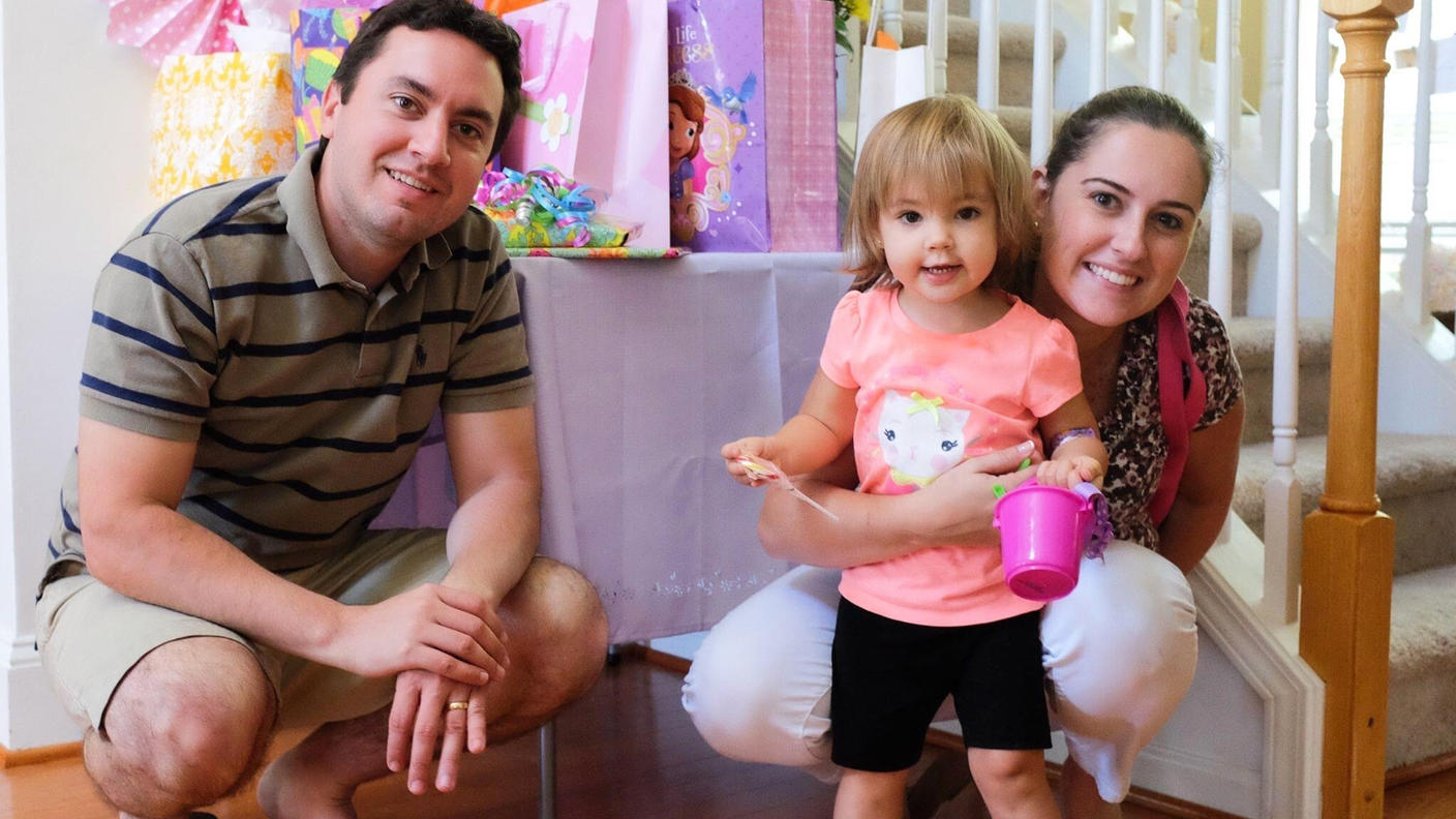 Joe and Erika smile and crouch next to their daughter, Melissa. Erika’s arm is around Melissa, who holds a small plastic bucket and smiles at the camera.