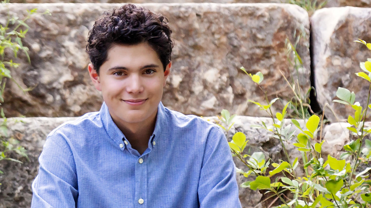 A young man, Callan, with brown hair and eyes, sits on a stone step wearing a blue button-up shirt and dress pants and smiles at the camera.