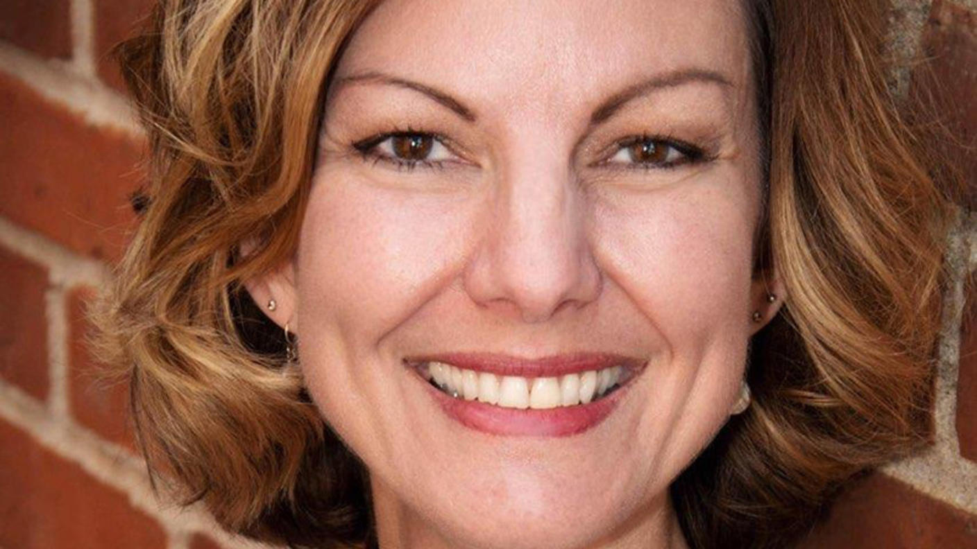 Woman with short, brown hair wearing a blue and white top smiling at the camera while leaning against a brick wall.