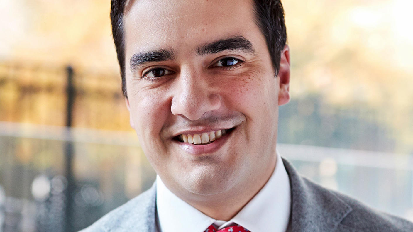 A man, Dr. Michael Ortiz, with brown hair and brown eyes and wearing a grey suit with a red tie, smiles at the camera.