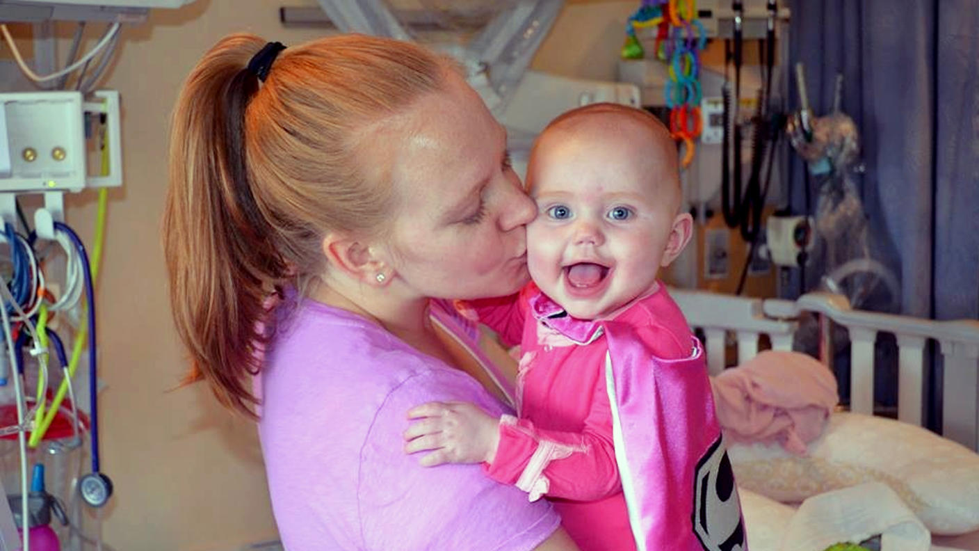 A woman, Kallie, with blond hair in a ponytail kisses the cheek of a smiling, blue-eyed baby, Sophie, wearing a pink superhero cape and facing the camera.