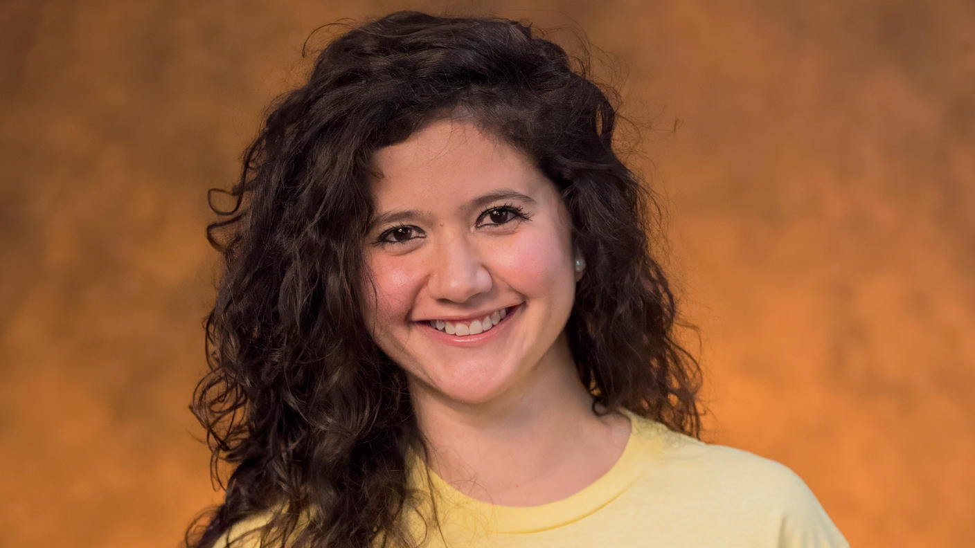Juanita stands smiling at the camera, with dark brown, wavy hair and dark brown eyes, wearing a yellow T-shirt printed with “Behold Be Gold.”