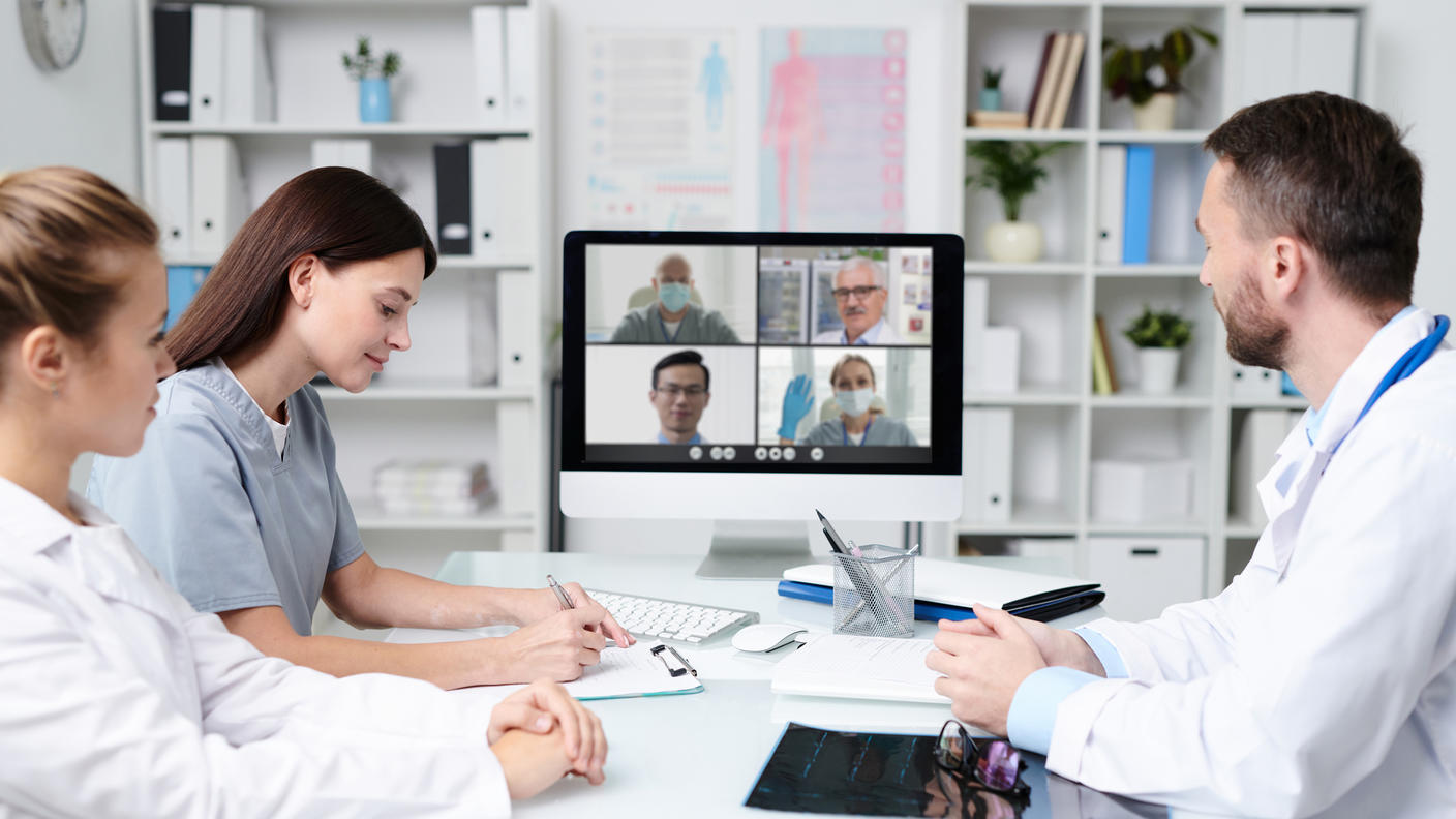 Three health care providers sit around a conference table having a videoconference with four health care providers working remotely. 