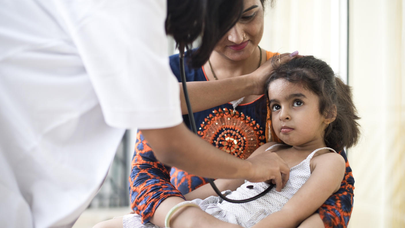 Una médica apoya un estetoscopio en el pecho de una niña que está en brazos de la madre. La niña está en brazos de la madre.