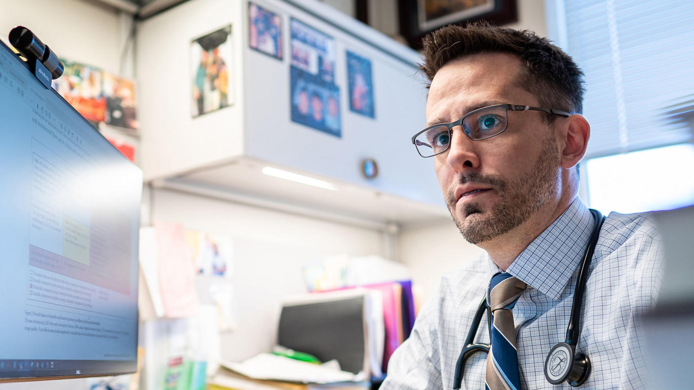 Dr. Matt Ehrhardt, with brown eyes and glasses, sits at a desk, pen in hand, wearing a dress shirt and tie and looking pensively into the distance.