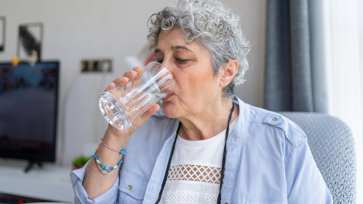 A person with short gray hair drinks water from a tall glass.