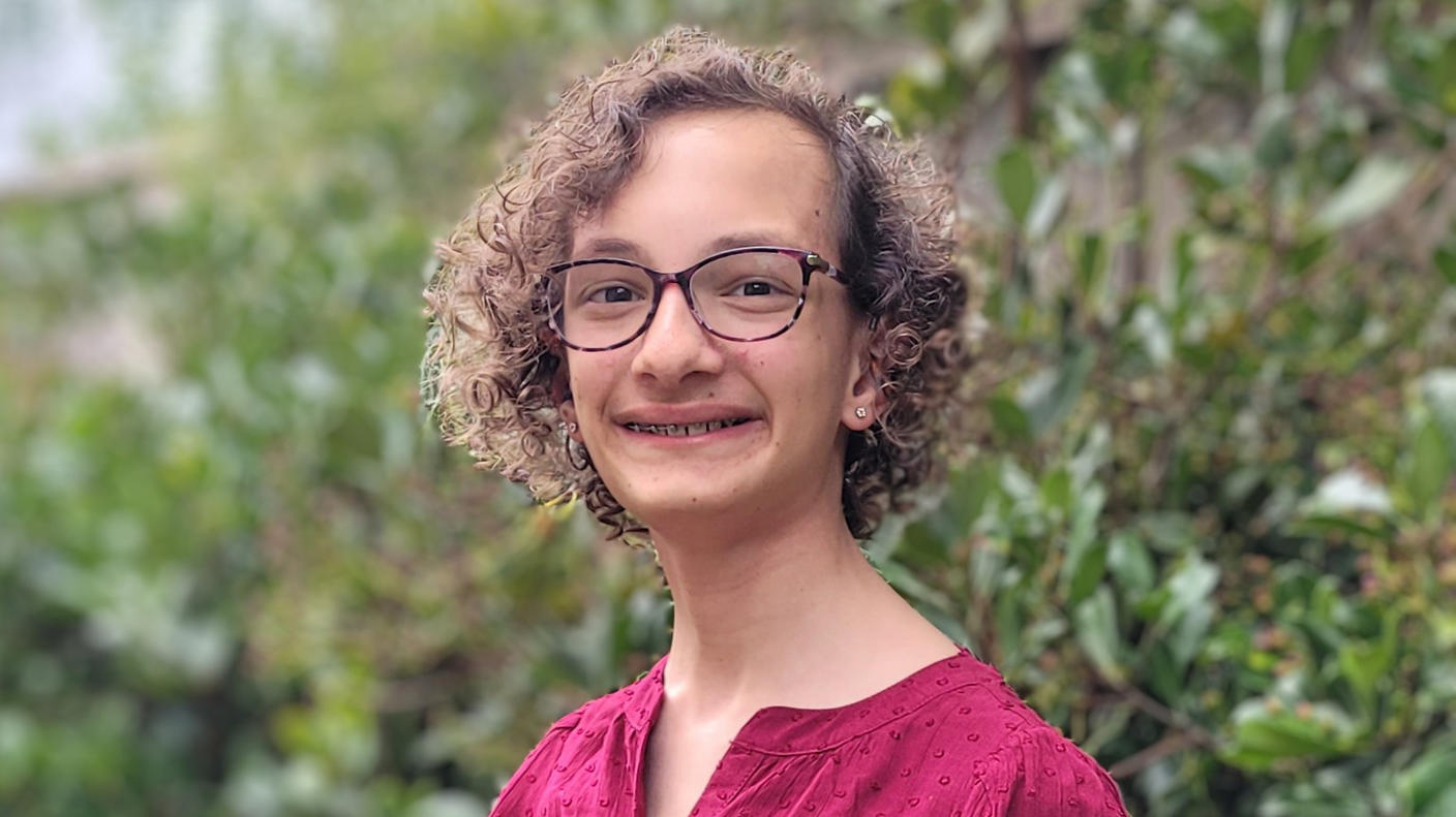 Young girl with curly hair, glasses, and a red dress smiling at the camera.