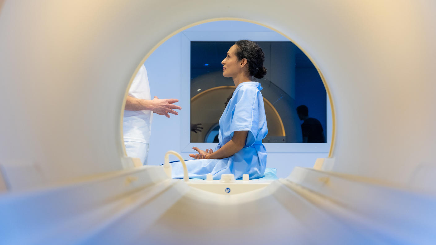 A woman viewed through the opening of an MRI machine. She is seated, wearing a hospital gown, and listening to a health provider who is standing in front of her.