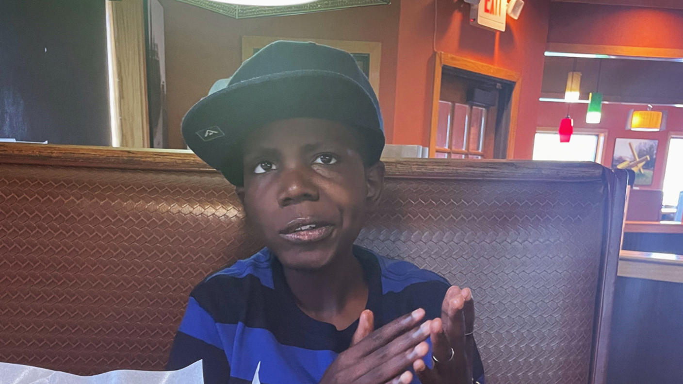 A boy, Cameron, wearing a ball cap and a black and blue striped T-shirt, sits in a restaurant booth and looks toward the camera.