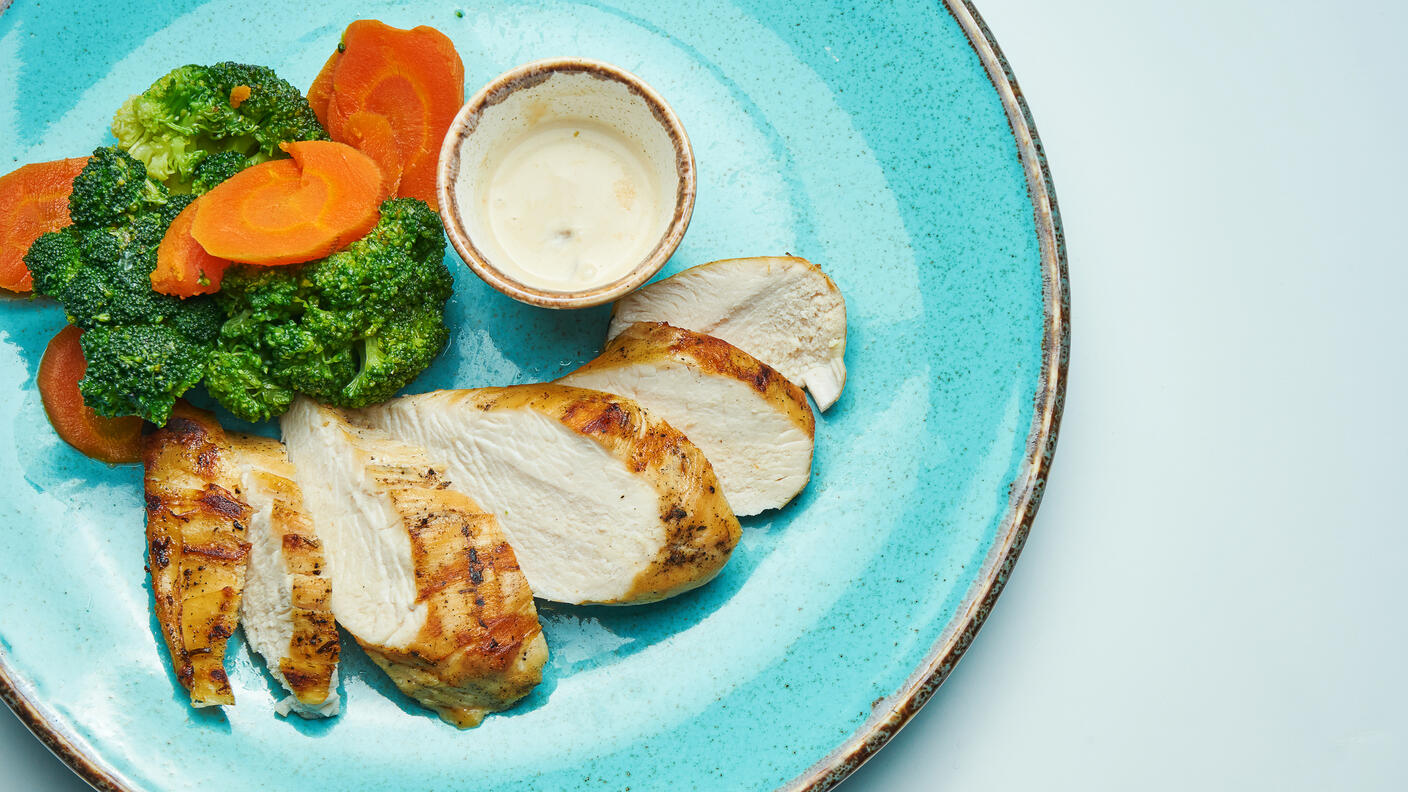 Slices of roasted chicken breast and cooked broccoli and carrots arranged on a blue plate along with a dip in a small bowl.