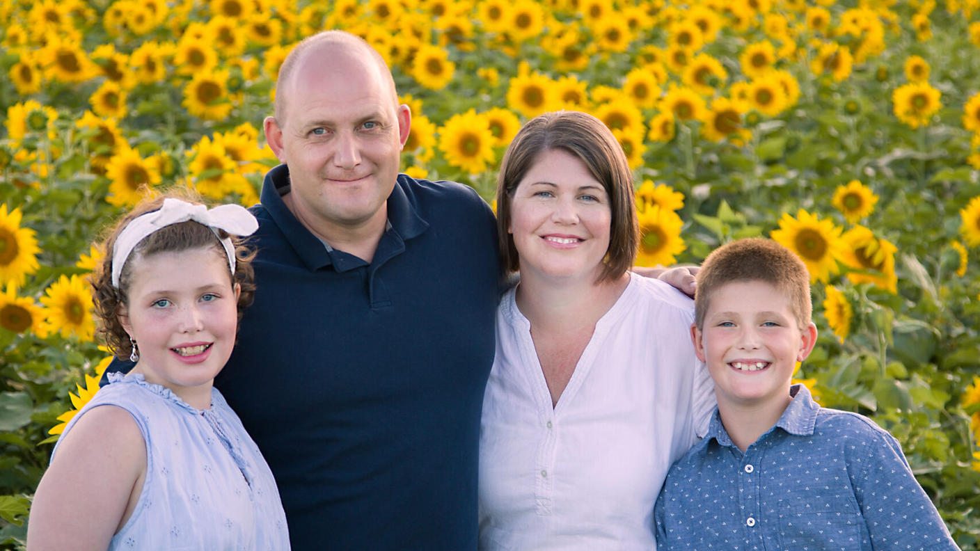 Girl in a blue dress with short hair held back with a headband, standing in a sunflower field with her family.