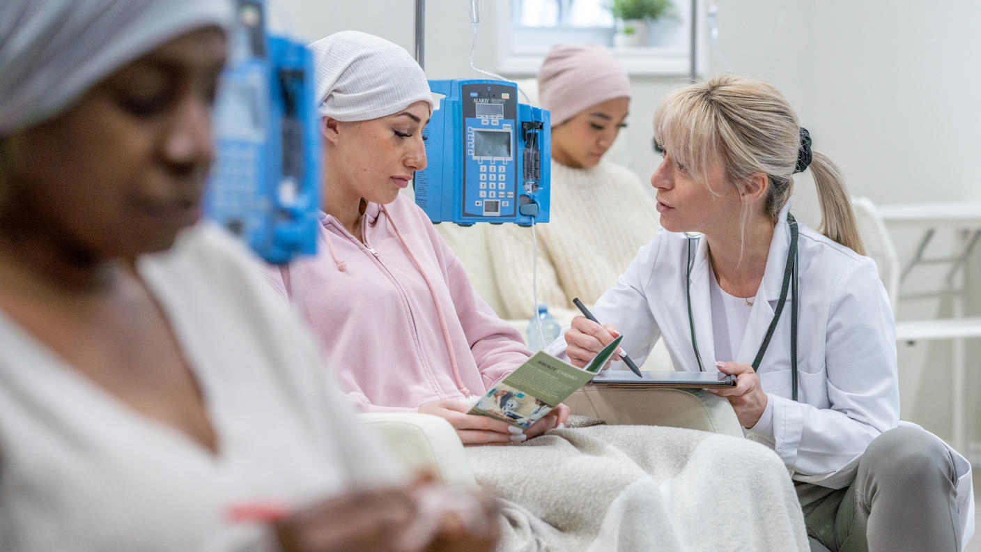 Three women sit in a clinic having IV infusions. A health provider kneels beside and chats with one young woman who is looking at a pamphlet.