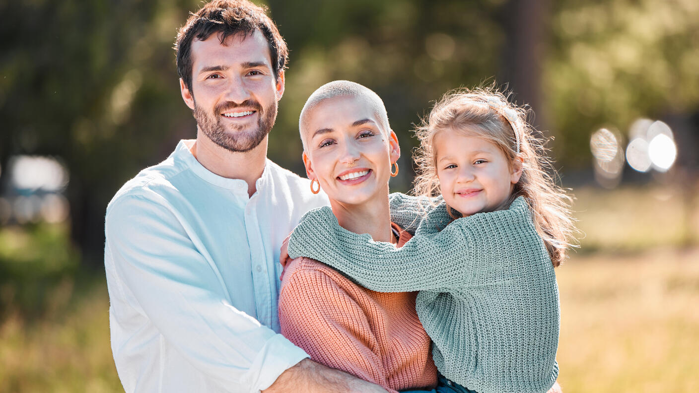 Cancer survivor holding young daughter in arms with husband hugging them both