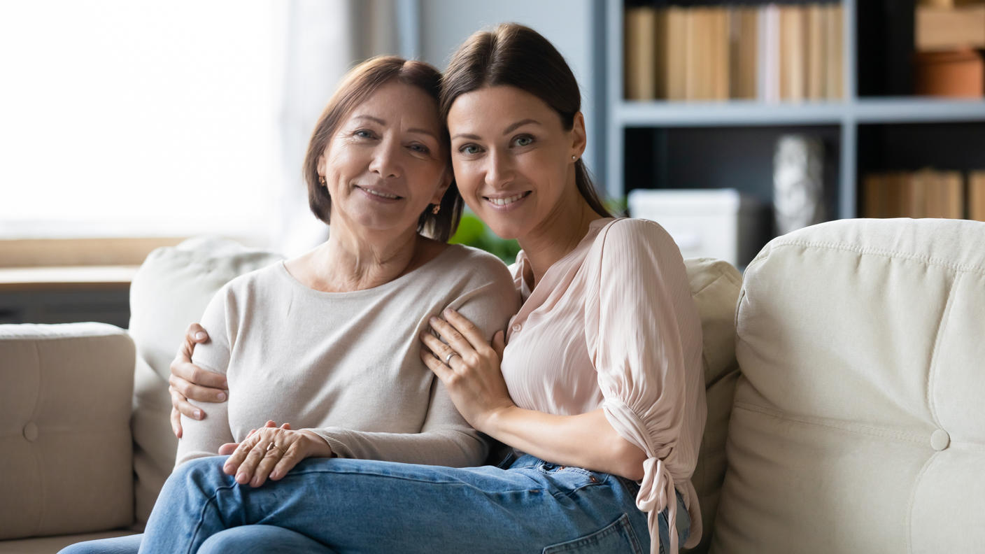 A smiling woman with her arm around an older smiling woman sitting together on a sofa.