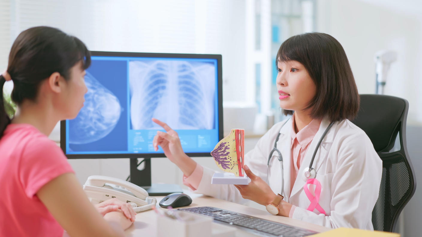A doctor holding a model of a breast and pointing at an image of a mammogram on a monitor while talking to a patient.