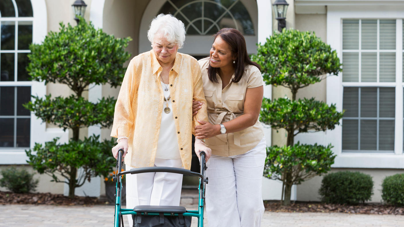 Senior woman with walker strolling with middle-aged woman helping her.