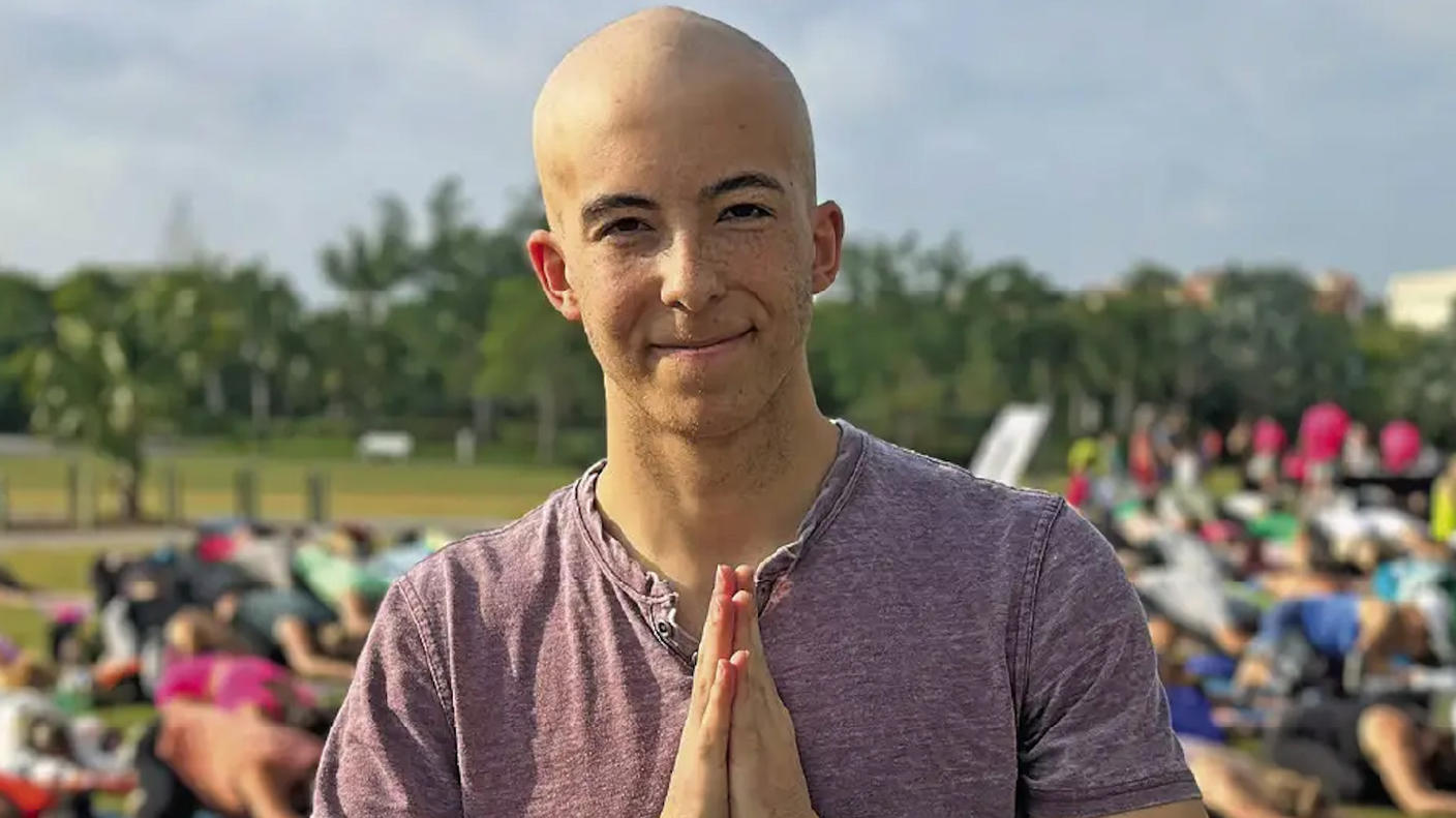 Jace, bald, smiling slightly, hands pressed together in prayer, stands outside in a crowd of people doing yoga.