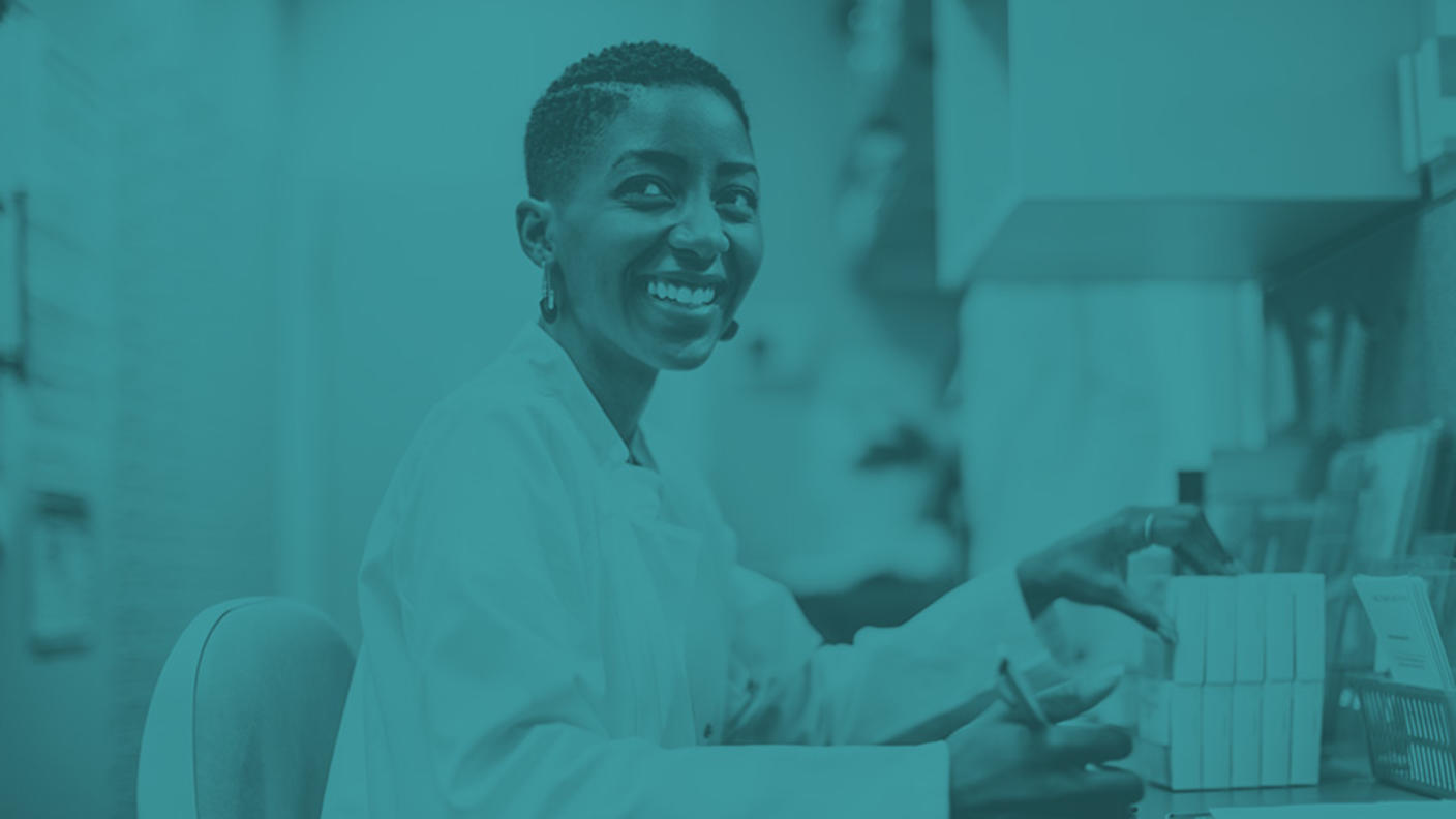 Woman wearing white labcoat is handling medication boxes while holding a pen and a clipboard.