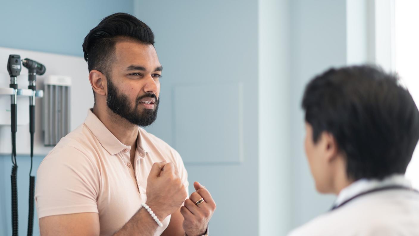 A young man with dark black hair and beard, form fitting orange shirt, with his hands in front of him speaking with a doctor. 
