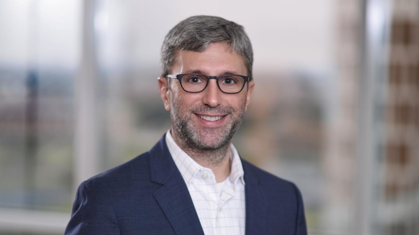 Man with grey and dark hair wearing glasses, a blue suit jacket, and a white dress shirt.