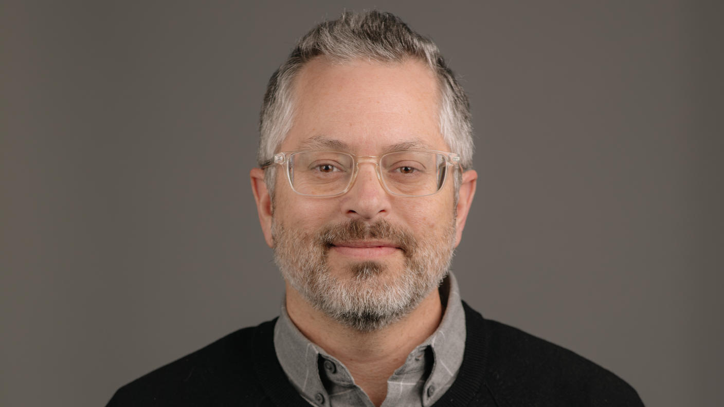 Bearded man wearing clear glasses and a black sweater over a grey collared shirt