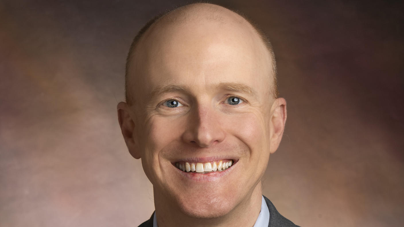 Man in grey suit, light blue shirt, and red, dotted tie smiles at the camera