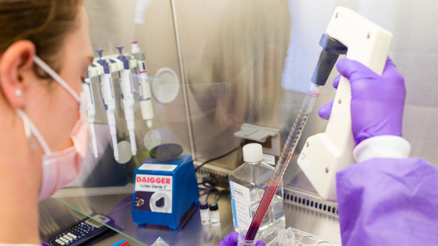 Health care provider in protective equipment uses a pipette to transfer red liquid into a laboratory tube inside a biosafety cabinet.