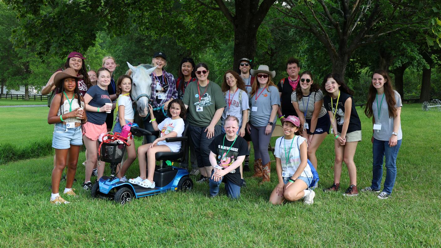 A large group of Camp Fantastic participants poses outdoors with a white horse under leafy trees, with one person seated on a mobility scooter.
