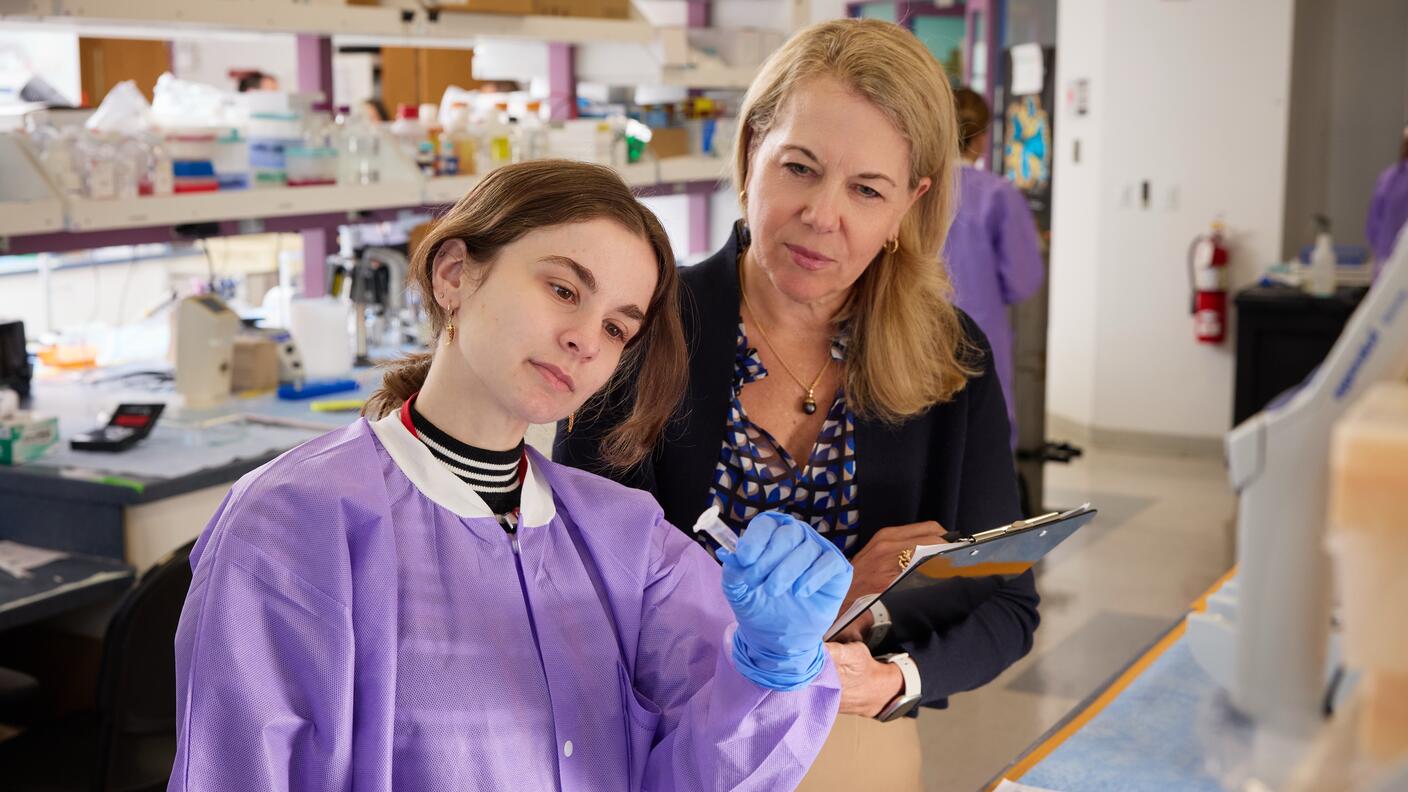 A laboratory researcher examines a sample while a colleague observes and takes notes.