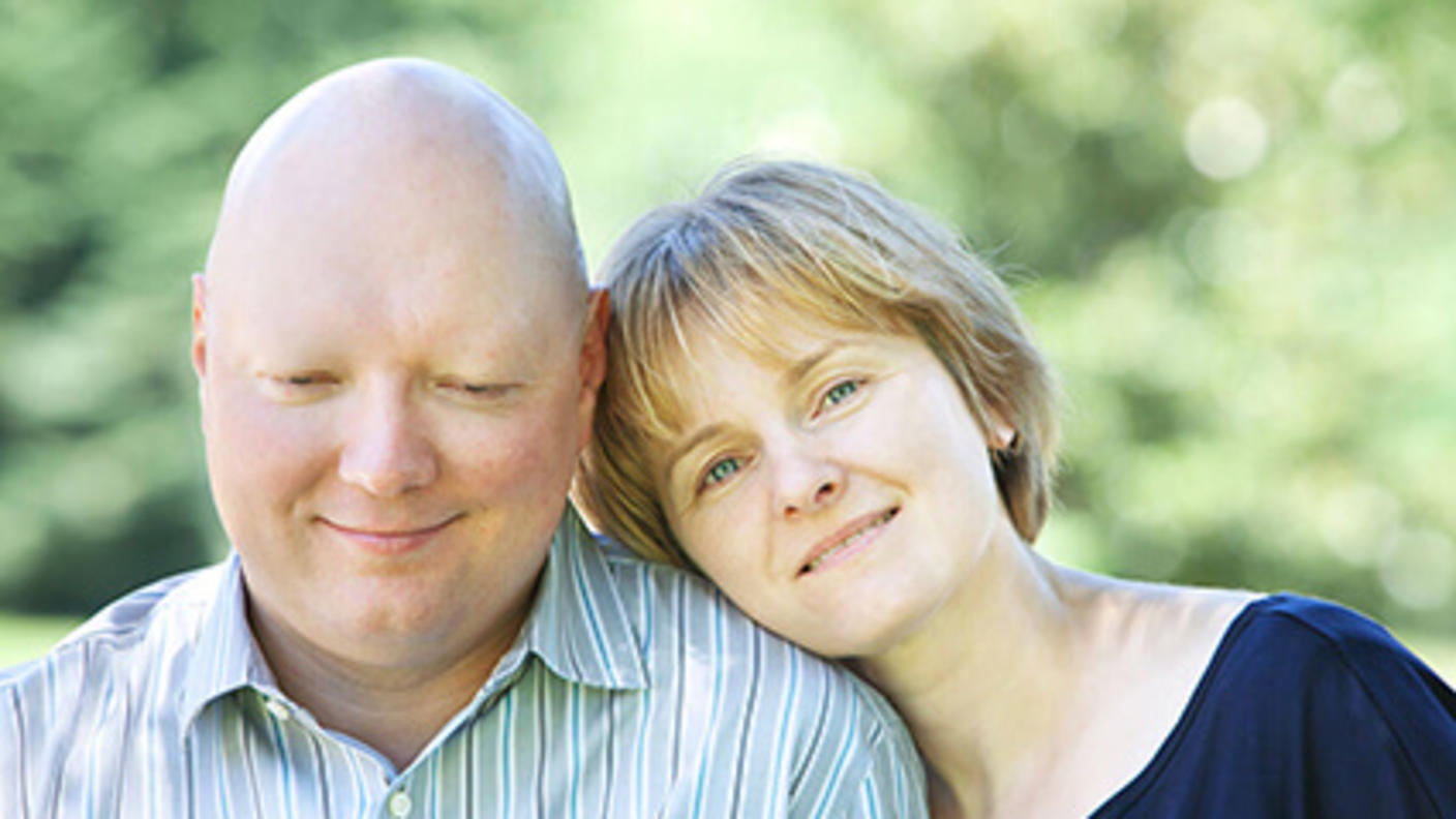 Man without hair smiles slightly as smiling woman rests her head on his shoulder.