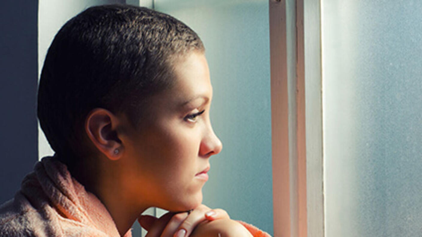A young woman with short hair and wearing a pink robe looks pensively out of a fogged window. The length of her hair is indicative of her having done chemotherapy recently, resulting in some hair loss.