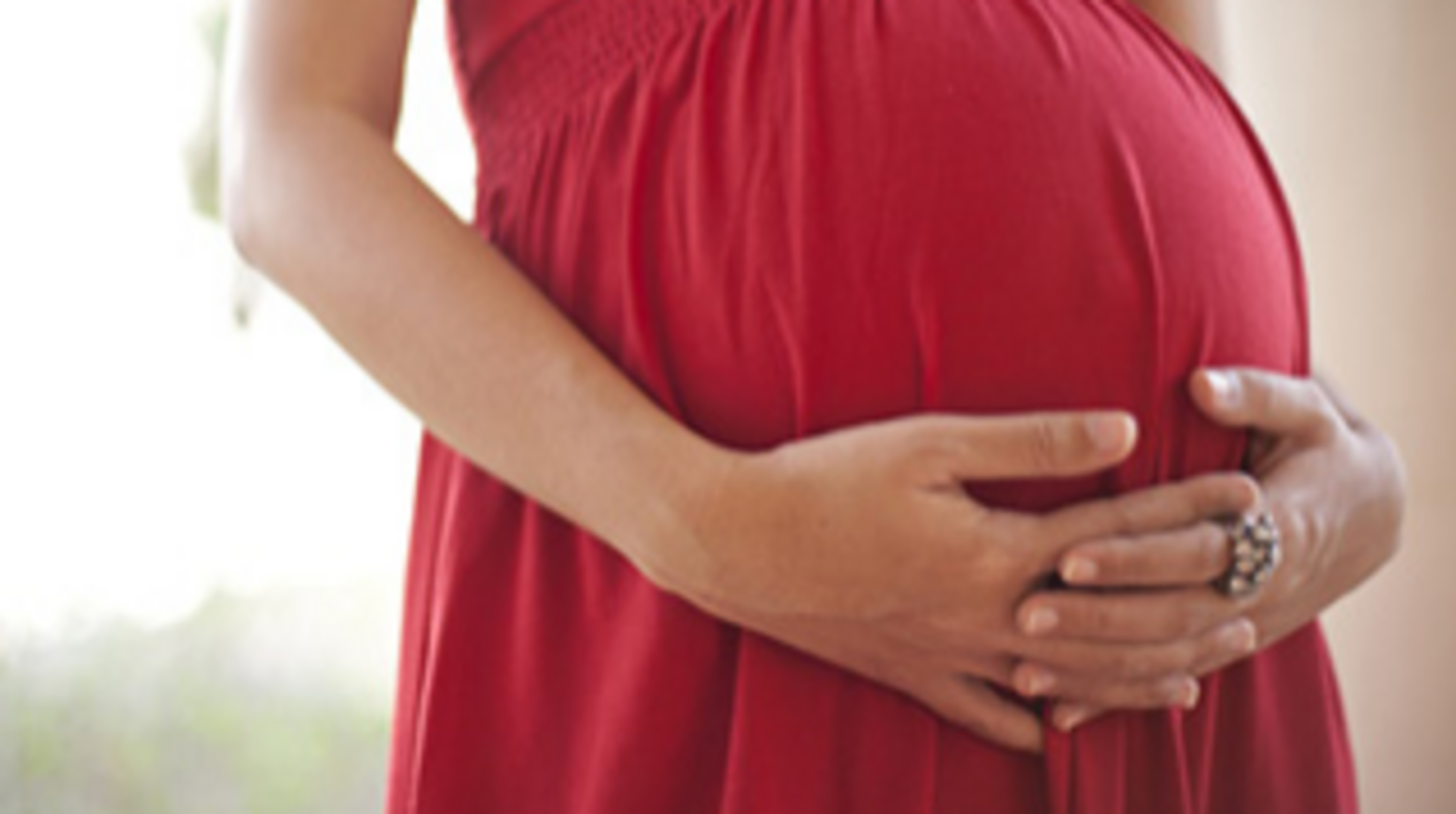 A close-up of a woman’s arms holding her pregnant belly. She is wearing a red dress.