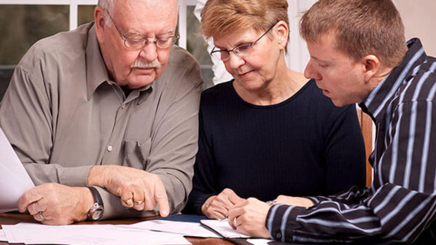 An elderly couple look down at papers on a table with a younger man. The elderly man is bald with a mustache and wears glasses with a button up shirt. The woman has short dirty blonde hair with glasses and a dark top. The younger man has short light brown hair and a striped button up shirt.