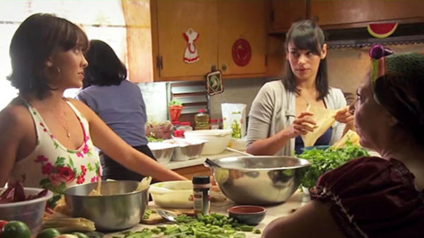 Two younger women in casual clothing look at each other while they prepare food. In the foreground, and also in shadow, an older woman looks on. Another woman is in the background with her back to the camera. The kitchen island in front of the two younger women and older woman has food and mixing bowls on it.