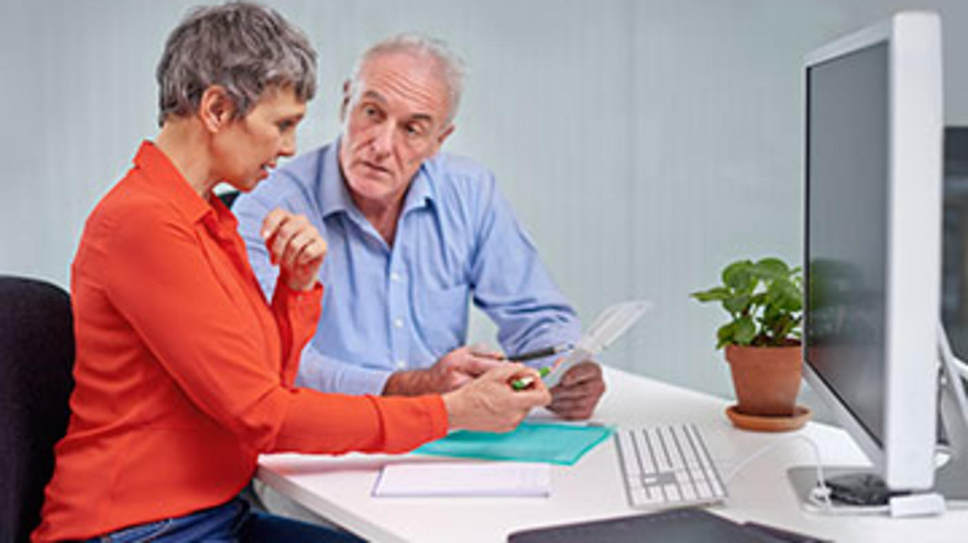 Woman and man seated in front of desktop computer and viewing paperwork.