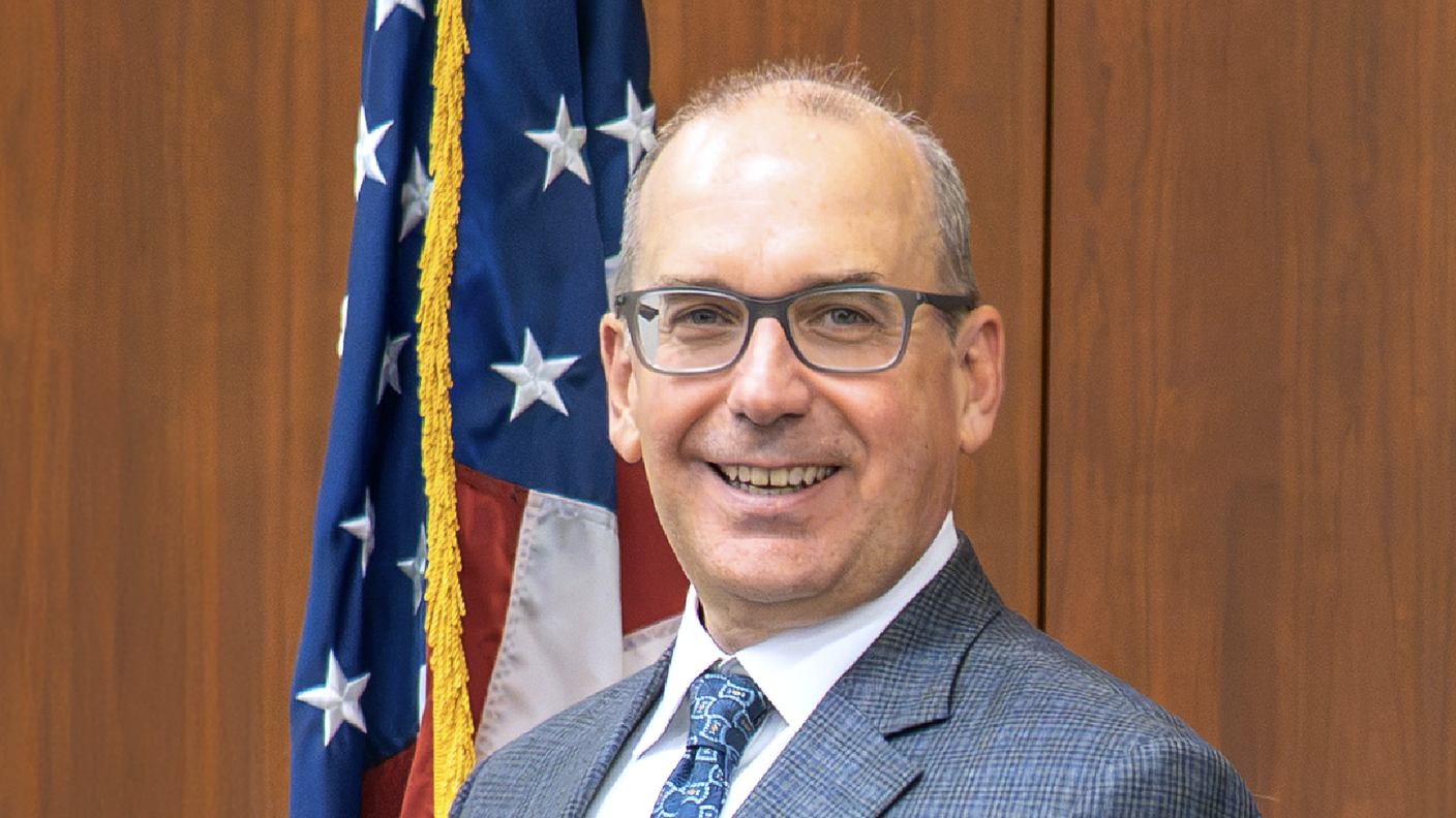 NCI Director Dr. Letai standing in front of a flag and brown wood background.
