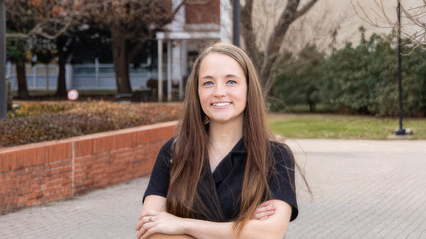 Gabrielle smiles at the camera with her arms crossed, wearing a black top and standing near a brick building and large tree.