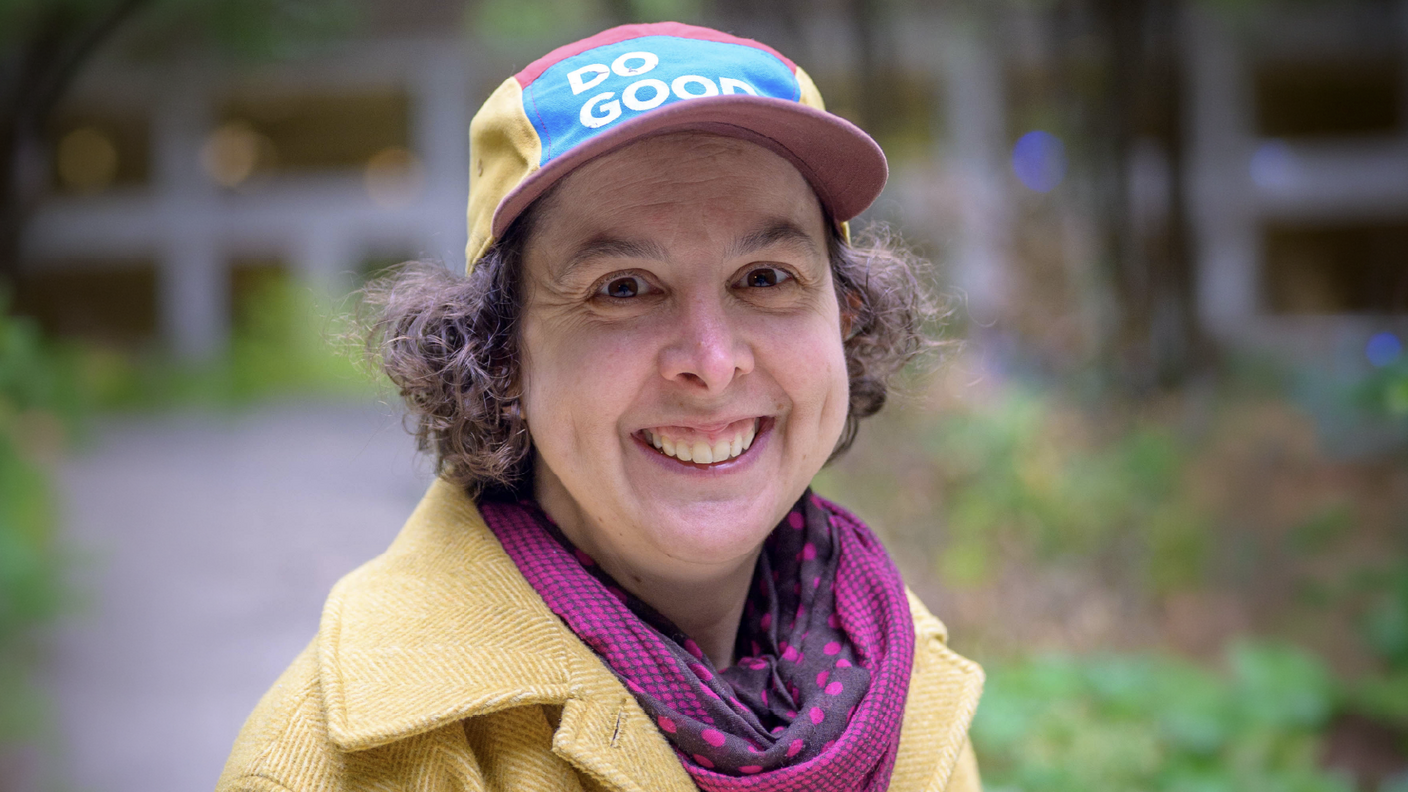 Olivia smiles at the camera, wearing a yellow jacket, patterned scarf, and multicolored cap with the words “DO GOOD.”