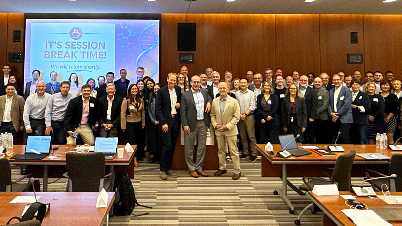 A large group of workshop attendees poses for a photo in a large meeting room. A screen behind them reads, “It’s session break time! We will return shortly.”